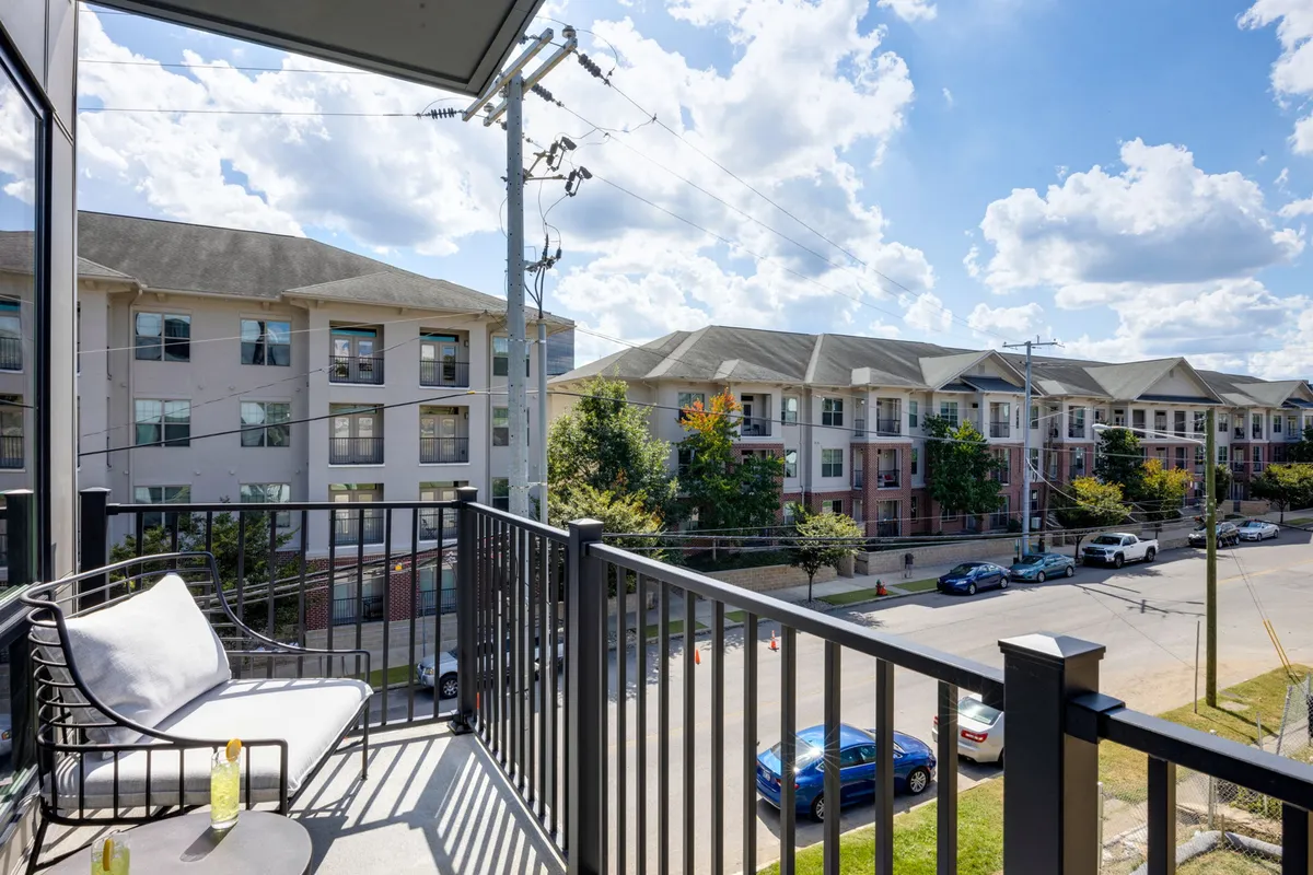The image presents an exterior view from the private balcony, with comfortable seating, a street view of neighboring buildings, and morning light filtering through trees. The mood is calm and welcoming, ideal for enjoying coffee or sunset cocktails while overlooking the city’s energy from a serene vantage point.