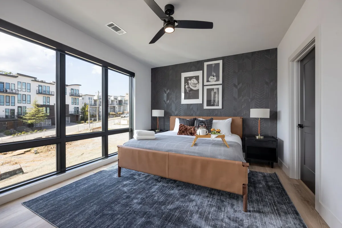 A spacious, sunlit bedroom featuring a large bed with a leather headboard, decorative wall art, and a nightstand with a lamp. The room feels calm and contemporary, with expansive windows letting in ample natural light—ideal for unwinding after a day in Nashville.
