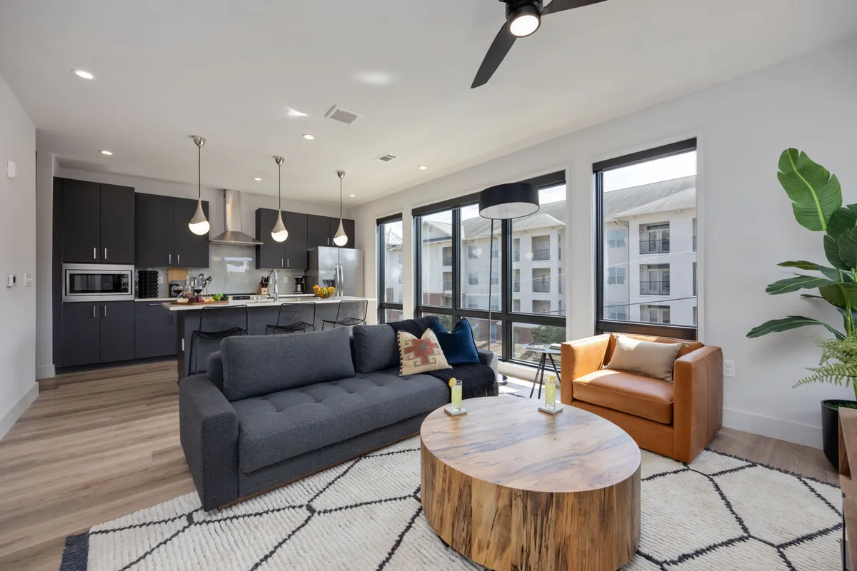 This image captures a cozy and modern interior living space featuring a plush sectional sofa, coffee table, kitchen area with sleek surfaces, large floor-to-ceiling windows allowing sunlight to flood the room, and a green plant adding a fresh touch. The open-concept design is perfectly set up for socializing, relaxing, or enjoying a sunset through the windows.