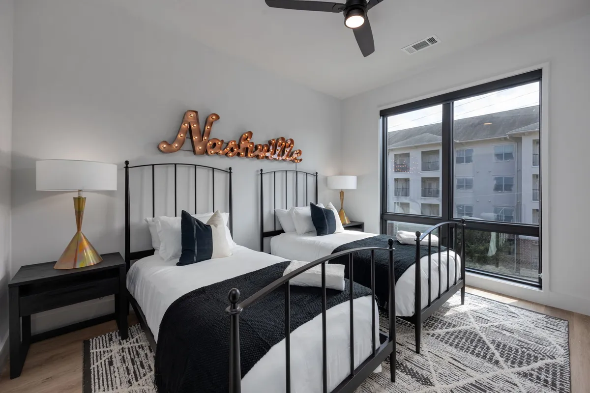 A bright bedroom featuring twin beds with black metal frames, a playful 'Nashville' sign, and a large window that fills the room with light. The decor is inviting and practical, making it ideal for families or friends sharing the space.