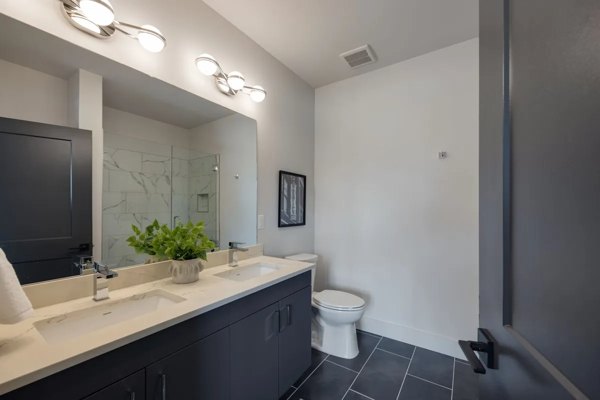 A modern bathroom with dual sinks, a large mirror, stylish light fixtures, and a green plant adding a touch of freshness. The space is bright and serene, designed for personal grooming and self-care.