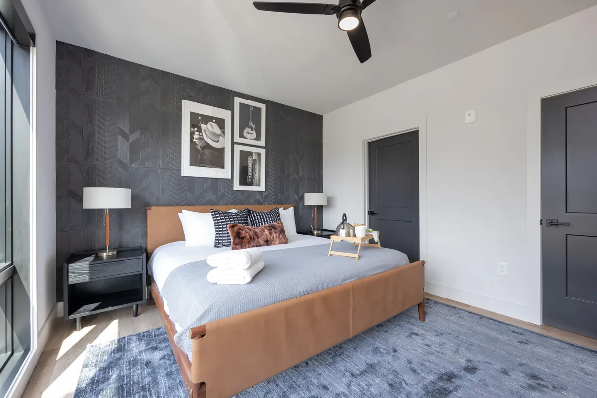 A cozy bedroom showing a bed with a brown leather frame, soft decorative pillows, and framed art on the wall. A nightstand with a lamp offers additional comfort, and the room’s palette exudes warmth and tranquility—perfect for relaxing after a long day.